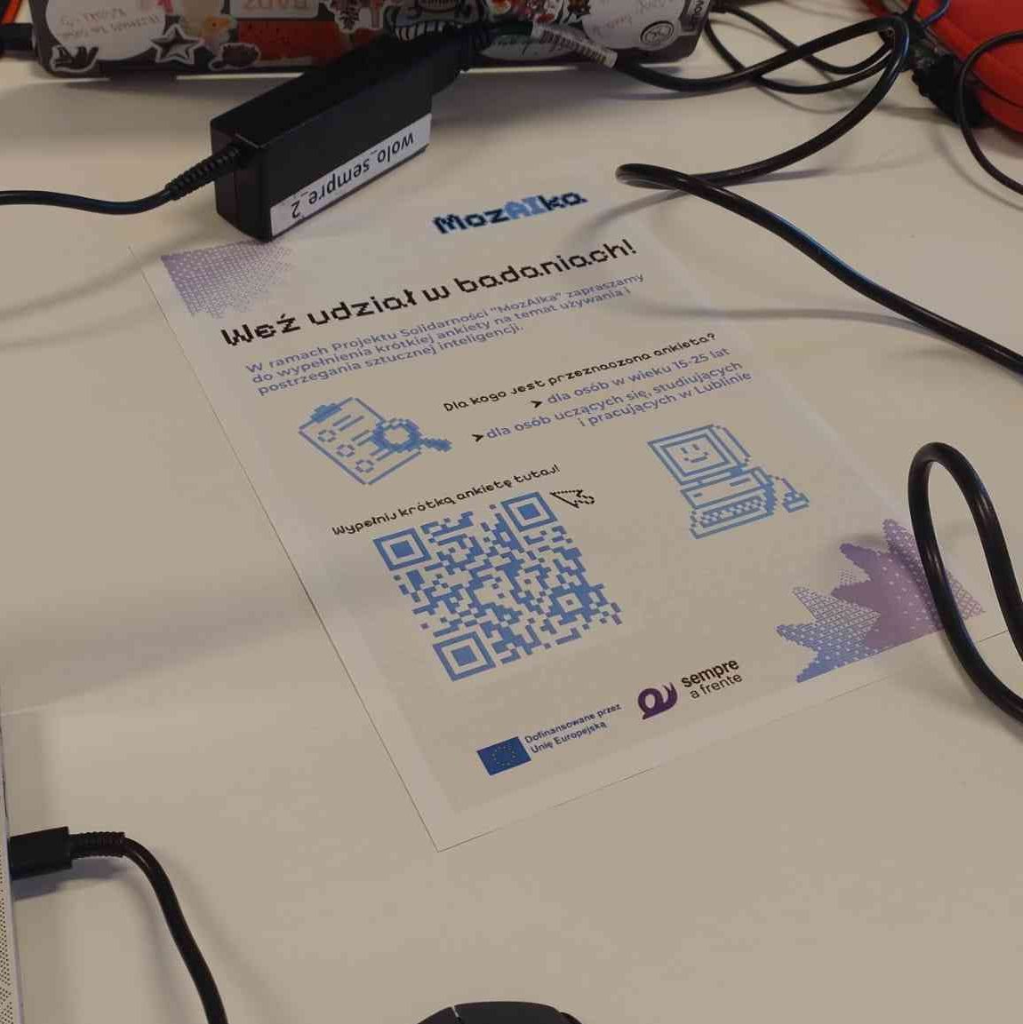 Ujęcie plakatu promującego udział w badaniu projektu MozAIka wśród kabli od laptopów