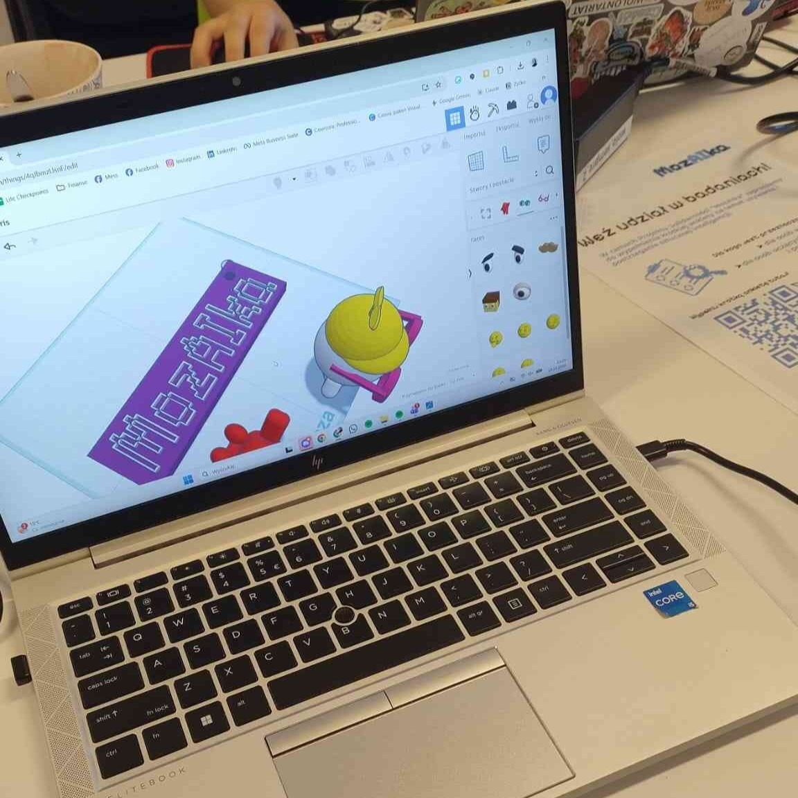 Ujęcie laptopa, na którym tworzone są modele 3D - widoczne jest logo MozAIki i widoczny od góry ludek w czapce i w okularach