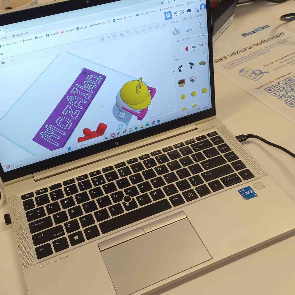 Ujęcie laptopa, na którym tworzone są modele 3D - widoczne jest logo MozAIki i widoczny od góry ludek w czapce i w okularach