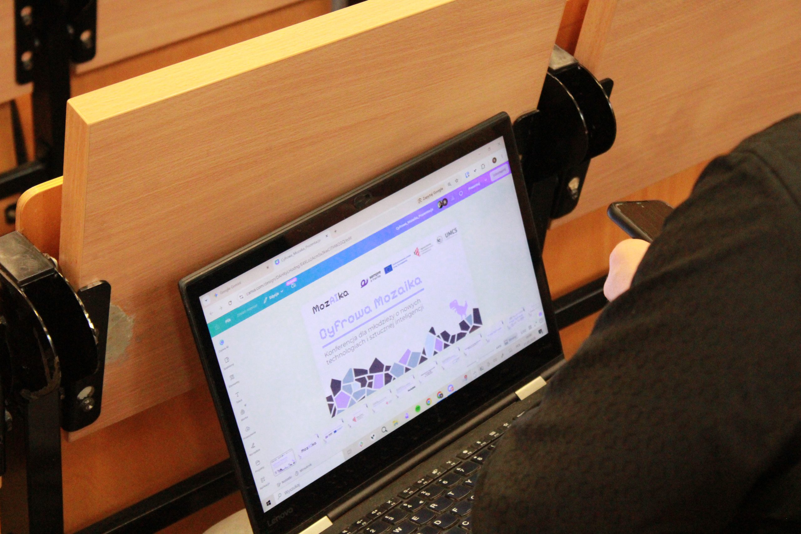 Ujęcie laptopa na kolanach niewidocznej osoby, na której widnieje grafika promocyjna konferencji "Cyfrowa MozAIka"