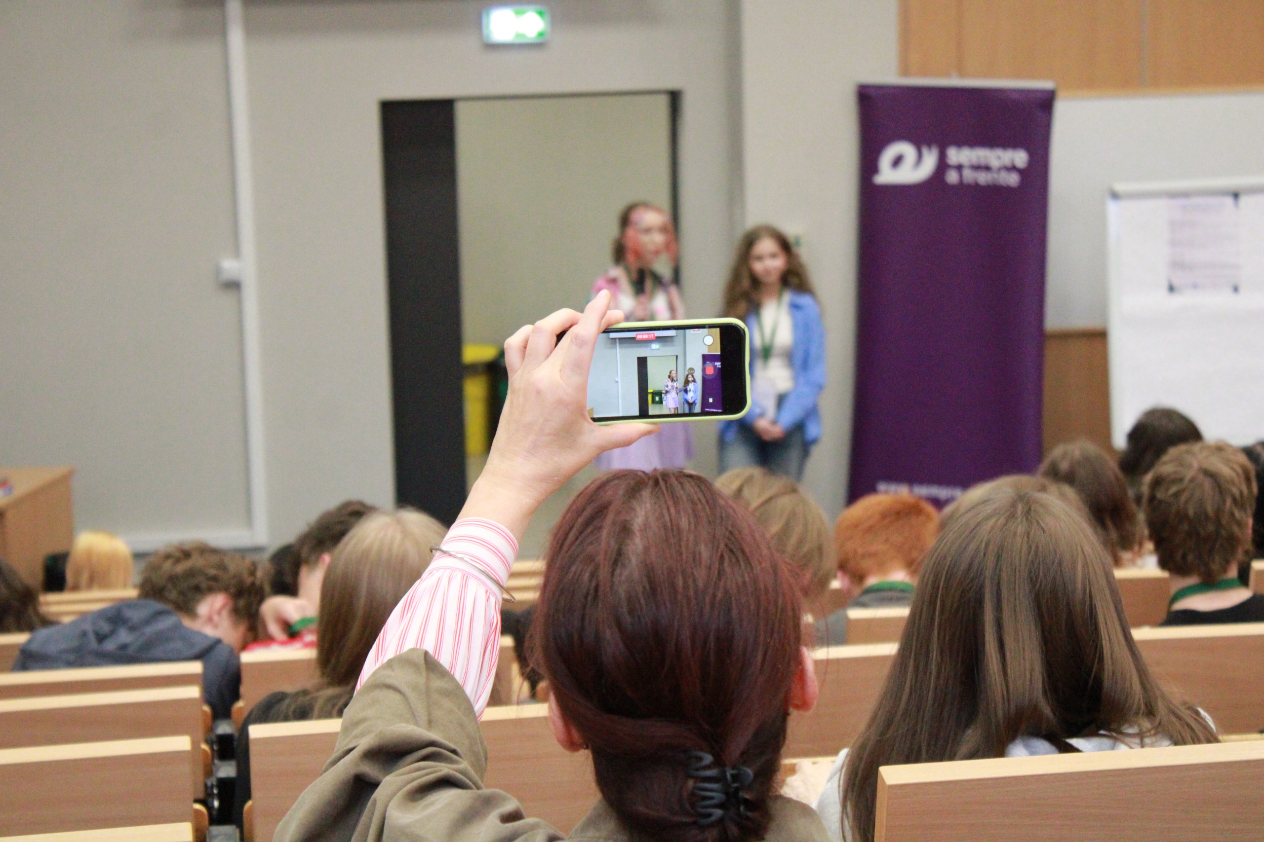 Osoba wśród siedzących uczestników na auli nagrywająca na telefonie wystąpienie dwóch uczestniczek podczas konferencji, z daleka można dostrzec baner roll-up Fundacji Sempre a Frente